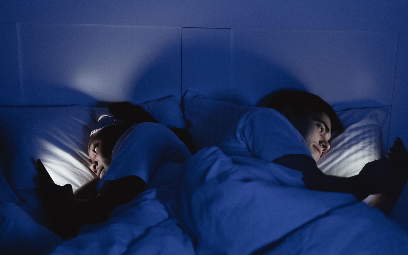 Two people lying in bed at night lit by the blue glow of their phone screens